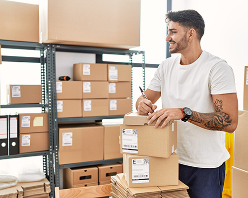 Amazon seller dressed in white tshirt and jeans writing on shipping box, boxes on metal shelf in background